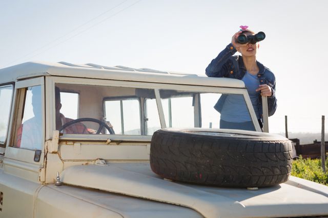 Woman standing next to a vehicle, looking through binoculars at the countryside on a sunny day. Ideal for themes related to adventure, travel, exploration, nature, and outdoor activities. Can be used in travel blogs, adventure magazines, and promotional materials for outdoor gear.