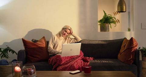 Cozy evening remote work scene woman on sofa with laptop wrapped in red blanket