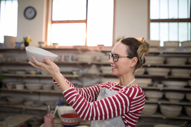Female potter wearing glasses and striped shirt inspecting a ceramic bowl in a pottery workshop. Shelves filled with pottery in the background. Ideal for use in articles about pottery, craftsmanship, creative arts, small businesses, and artisan workshops.