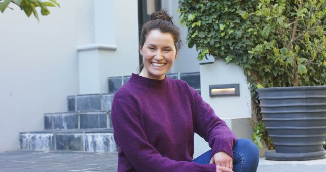 Smiling woman sitting in front of home entrance, exuding a casual and relaxed vibe. She is wearing a purple sweater and jeans, adding a cozy touch. Ideal for advertisements related to real estate, home decor, or everyday life and happiness scenes.