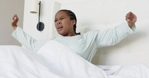 African american woman stretching and yawning while sitting up in bed wearing mint pajamas