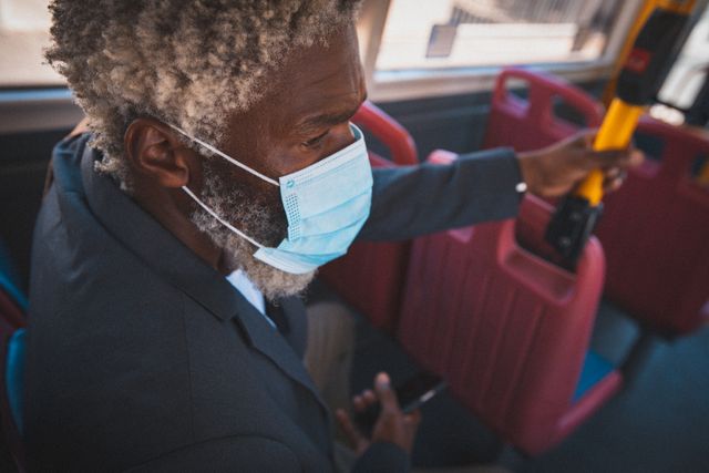 African american senior man wearing face mask sitting in public bus holding smartphone. digital nomad on the go in the city during coronavirus covid 19 pandemic.
