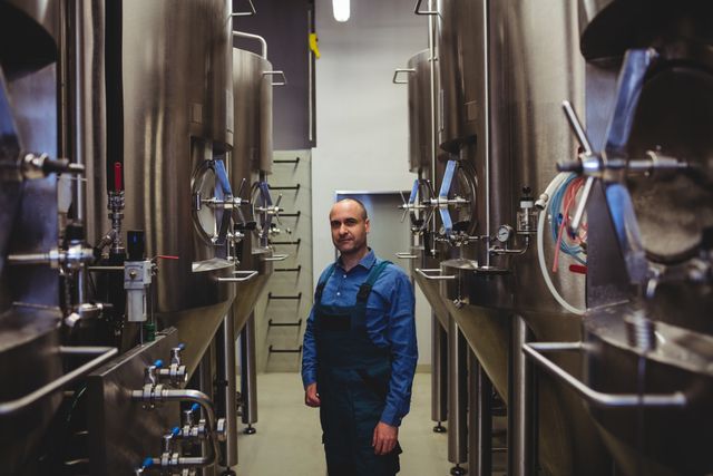 Confident brewer standing amidst large stainless steel tanks and machinery in a brewery. Ideal for use in articles or advertisements related to brewing, manufacturing, industrial work environments, and professional craftsmanship. Can be used to depict the brewing process, industrial settings, or professional expertise in the beverage industry.