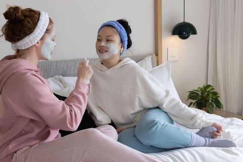 Multicultural friends applying facial masks on bed enjoying cozy at-home skincare moment
