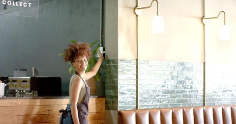 Smiling barista raising arm at modern cafe counter with green tile column and leather booth
