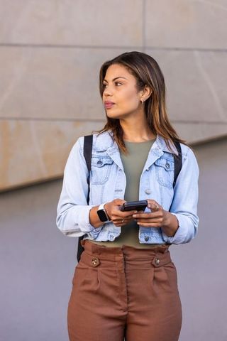 Young woman standing with smartphone in urban plaza, denim jacket backpack commuter