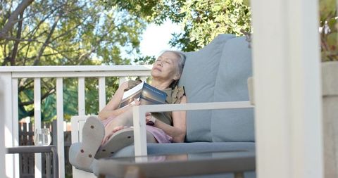 Senior asian woman relaxing on sunlit porch sofa reading paperback book, outdoor leisure