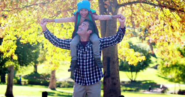 A father is carrying his daughter on his shoulders, enjoying quality time outdoors in a park during autumn. The trees in the background showcase fall foliage. Ideal for concepts related to family bonding, joy, parenting, outdoor activities, and seasonal sentiments.