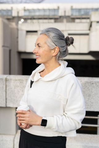 Stylish senior woman wearing white hoodie holding coffee and smartwatch on urban rooftop
