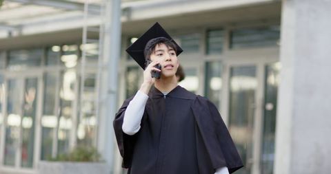 Asian graduate calling on smartphone outside modern campus entrance wearing cap and gown