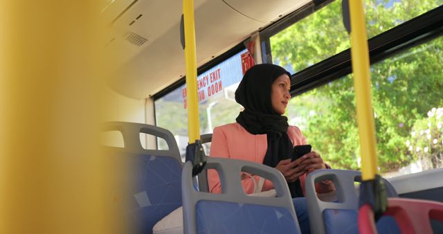 This stock photo shows a Muslim woman wearing a hijab and casual attire, sitting alone on a public bus and using a smartphone while looking out of the window. The scene captures a peaceful moment during daytime transportation. This image is ideal for illustrating concepts related to diverse city life, public transit usage, mobile technology, and personal reflection during commuting.