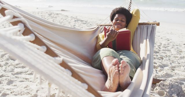 This image shows a woman lying comfortably on a hammock at a beach, reading a book. Perfect for promoting vacation packages, beach resorts, or leisure products. It depicts themes of relaxation, leisure, and personal enjoyment.