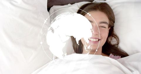 Smiling woman lying in bed with white duvet and digital globe overlay suggesting global connectivity