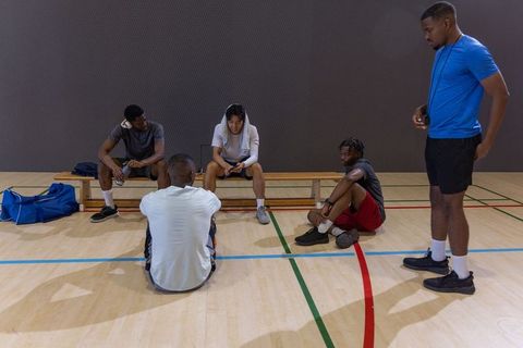 Multiethnic male basketball teammates resting and checking phone on indoor gym bench