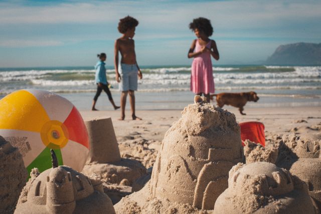 African American children are enjoying a sunny day at the beach, building sandcastles and playing with a beach ball. In the background, a dog and another person are walking along the shore. This image is perfect for promoting family vacations, summer activities, and outdoor fun. It can be used in travel brochures, family-oriented advertisements, and websites focusing on beach destinations.