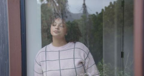 Thoughtful woman gazing through glass door with soft reflections and pastel grid sweater