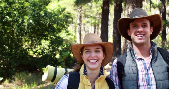 Happy couple hiking through a dense forest, carrying backpacks and wearing wide-brimmed hats. They both have big smiles, indicating enjoyment of an adventurous outdoor activity. Perfect for illustrating themes of outdoor leisure, travel, nature exploration, and active lifestyles.