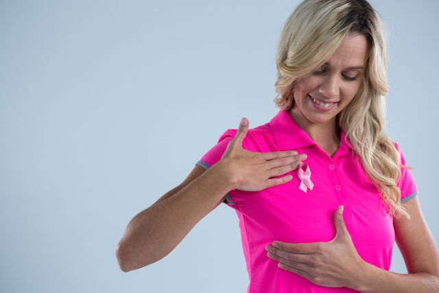 Woman wearing a pink ribbon on her shirt, symbolizing breast cancer awareness. She is smiling and touching her breast, promoting health and prevention. This image can be used for health campaigns, awareness programs, and support initiatives related to breast cancer.