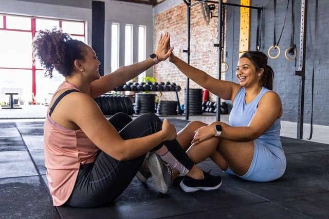 Two diverse women high-fiving during partner sit-up workout in bright industrial gym