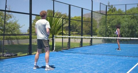 Playing padel on blue court, man and woman volleying across net under sunlit sky