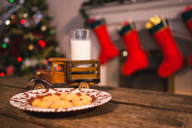 Christmas cookies and a glass of milk on a wooden table with a toy truck, set against a festive background featuring a decorated Christmas tree and stockings hanging by a fireplace. Ideal for holiday-themed promotions, greeting cards, festive blog posts, and social media content.