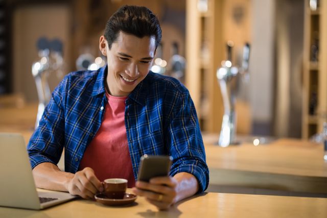 Young man smiling while using a mobile phone in a coffee shop. Ideal for illustrating modern lifestyle, communication, technology use in cafes, and casual social interactions. Perfect for marketing materials, social media content, or blog posts focusing on technology, relaxation, and coffee culture.