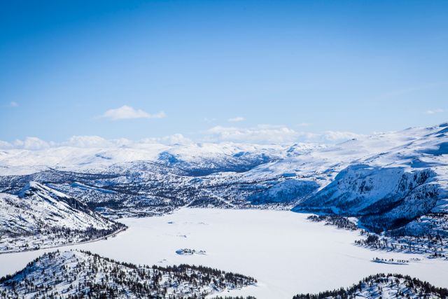 Aerial perspective showcasing stunning snowy mountains and an icy lake under a vibrant blue sky. Perfect for promoting winter tourism, travel destinations, nature-themed publications, outdoor adventure activities, and scenic backgrounds for presentations or websites.