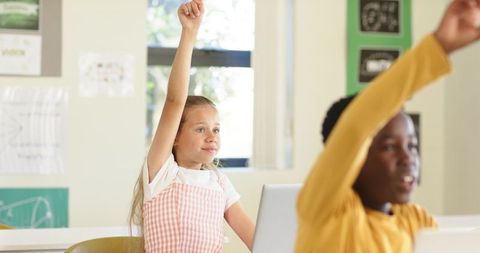 Diverse children raising hands in classroom with laptop and learning posters