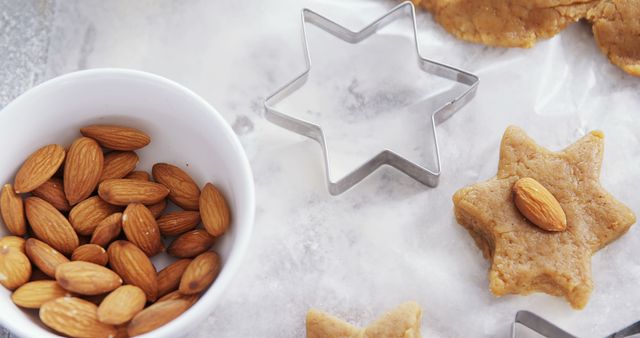 Almonds in a white bowl and a star-shaped cookie cutter on a marble countertop, with a star-shaped cookie beside it, with copy space. Ingredients and tools for baking are presented, suggesting someone is preparing homemade cookies.