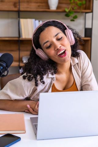 African american woman recording podcast at home with pink headphones and laptop