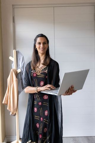Indian professional holding laptop in chic home office wearing black print dress portrait