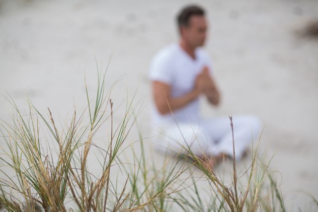 Mature man practicing meditation on a sandy beach with dune grass in the foreground. Suitable for wellness, mindfulness, and relaxation themes in promotional materials or articles. Can be used for promoting yoga, meditation retreats, and nature therapy.