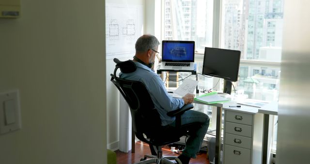 Male architect working on building plans at desk in modern office with city view. Could be used for themes related to architecture, urban planning, professional workplaces, or modern working environments.