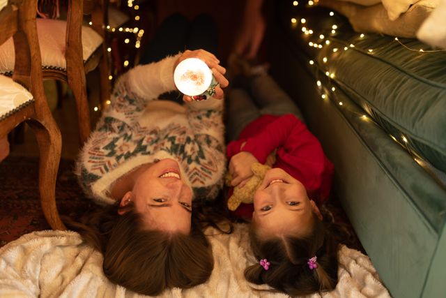 Mother and daughter lying on the floor in a cozy blanket fort, holding a glowing Christmas globe. The fort is illuminated with Christmas lights, creating a warm and festive atmosphere. Perfect for use in holiday-themed promotions, family bonding concepts, and winter celebration advertisements.
