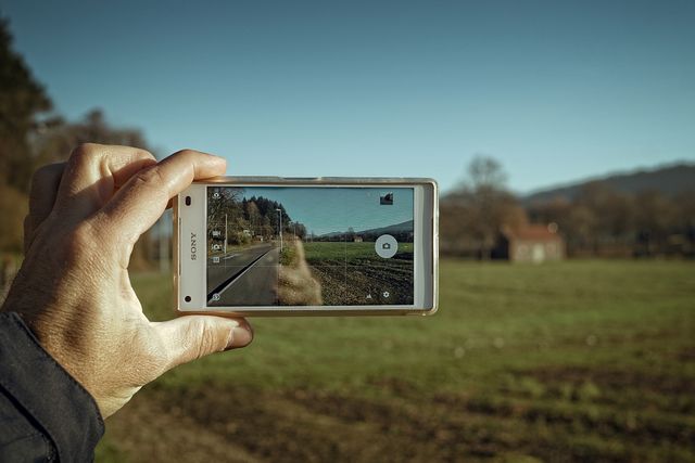 Hand holding smartphone, taking picture of scenic outdoor landscape with clear sky, open field, and distant building. Ideal for use in articles or advertisements related to smartphone photography, travel, outdoor activities, and nature appreciation.