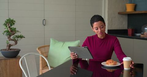 Mid-adult woman using tablet while reaching for coffee in cozy kitchen with croissant