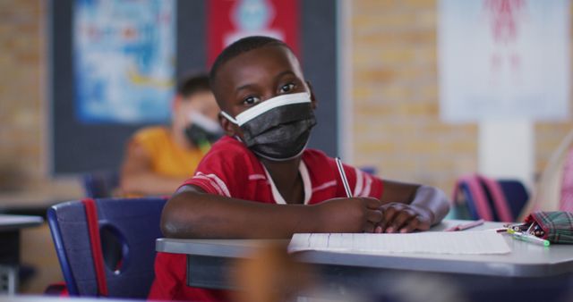 Portrait of african american schoolboy wearing face mask, sitting in classroom looking at camera. children in primary school during coronavirus covid 19 pandemic.