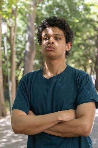 Pensive young african american man standing with crossed arms in sunlit tree-lined walkway