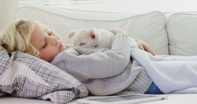 Blonde child resting on couch with white puppy on chest, displaying cozy, serene moment indoors. Ideal for themes of childhood, relaxation, bonding with pets, home comfort, and peaceful living. Useful for advertising family-friendly products, pet-related items, or home furnishings.