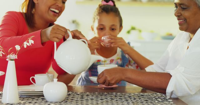 A multi-generational family enjoys a warm tea time together, with the young girl sipping from her cup as the adults pour and laugh. Capturing a moment of joy and connection, the image reflects a cozy, domestic atmosphere.