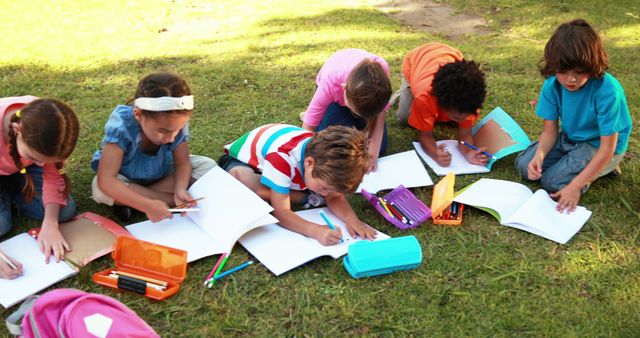 Children sitting on lawn drawing together in notebooks surrounded by pencils and art supplies. Suitable for illustrating concepts of outdoor learning, creativity in nature, group activities, and teamwork among children.