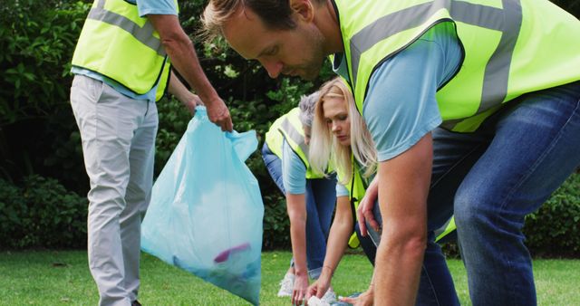 Group of volunteers wearing yellow vests cleaning up a park by picking up trash and litter from the grass with garbage bags. Useful for content on community service projects, environmental conservation efforts, and teamwork in public service activities.