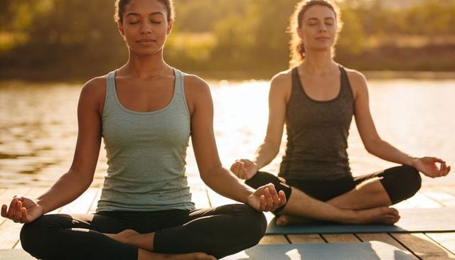 Black woman meditating on dock with companion at calm lake during golden hour
