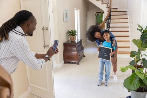 Parent photographing child holding 1st day of school letter board in bright home hallway