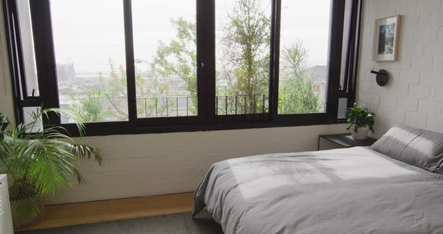 Image showcasing a modern bedroom interior with large windows that offer expansive city views. Room features a cozy bed with grey bedding, minimalistic decor, and several indoor plants enhancing a natural, serene atmosphere. Ideal for illustrating concepts related to urban living, modern interior design, and lifestyle blogs. Suitable for use in articles, real estate listings, home decor inspiration, and relaxation content.