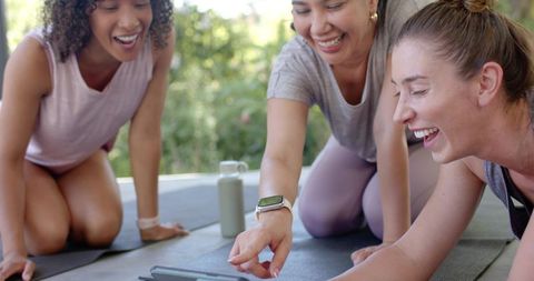Friends practicing yoga and laughing outdoors while using tablet for workout guidance