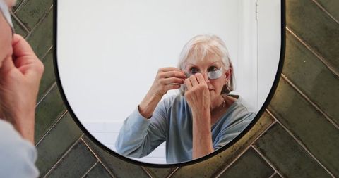 Senior woman adjusting under-eye patch in modern bathroom