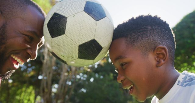 African american dad and son balancing the football together with their heads in the garden. fatherhood and love concept