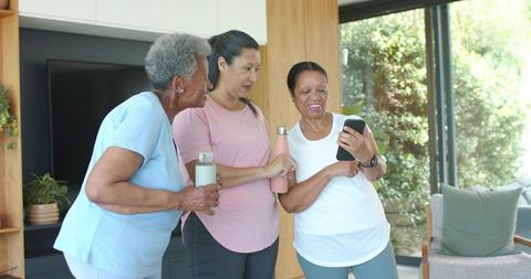 Senior women sharing fitness moment, diverse friends checking smartphone and hydrating in sunny home