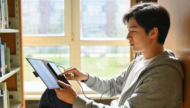Asian student charging phone with portable solar panel on sunny library window bench